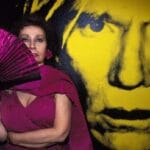 L'artista Ultraviolet con vestito e ventaglio viola è davanti a una grande foto di Andy Warhol