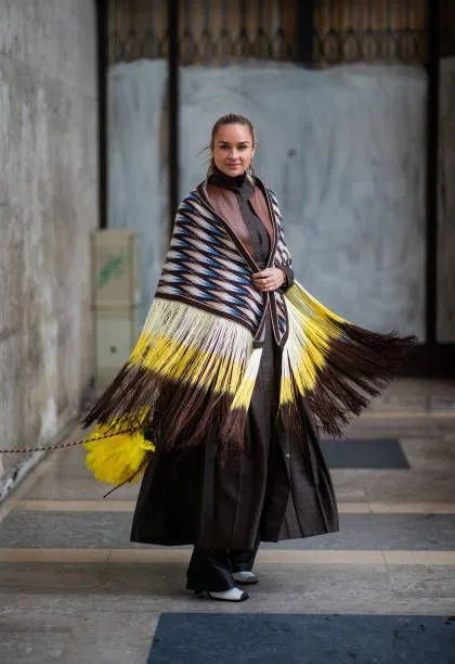 Donna indossa mantella a blocchi di colore giallo e marrone con lunghe frange sopra un trench marrone e pantaloni neri.