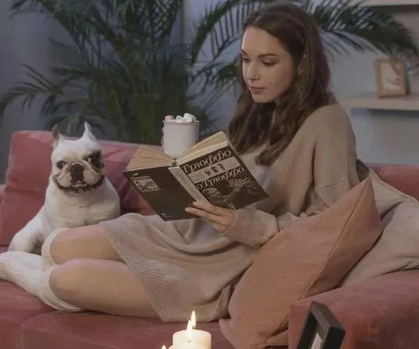 Una donna nella solitudine della sua casa si rilassa leggendo un libro sul divano con cuscini, cane e candele accese