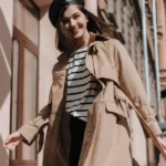 Ragazza sorridente con trench beige, maglietta a righe e berretto nero. Outfit casual chic da mezza stagione.