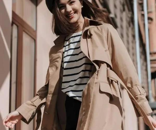 Ragazza sorridente con trench beige, maglietta a righe e berretto nero. Outfit casual chic da mezza stagione.