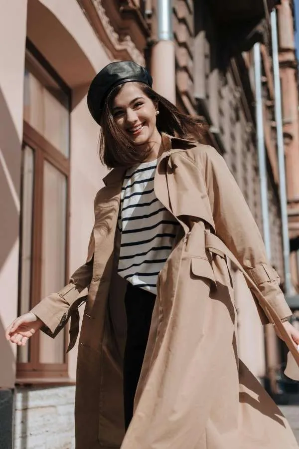 Ragazza sorridente con trench beige, maglietta a righe e berretto nero. Outfit casual chic da mezza stagione.