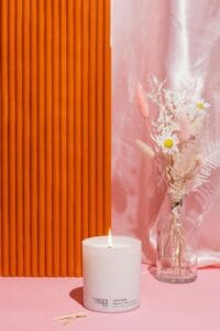 Una candela bianca accesa con accanto un vasetto di fiori secchi e margherite su sfondo rosa e arancione, idea regalo festa della mamma per Nice to be Woman