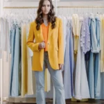 Ragazza indossa un outfit con blazer oversize giallo, top arancione e jeans chiari davanti a un appendiabiti, per la guida su come indossare i capi del guardaroba femminile.