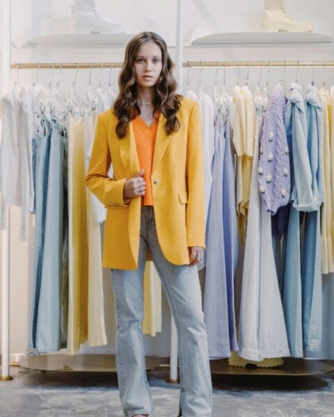 Ragazza indossa un outfit con blazer oversize giallo, top arancione e jeans chiari davanti a un appendiabiti, per la guida su come indossare i capi del guardaroba femminile.