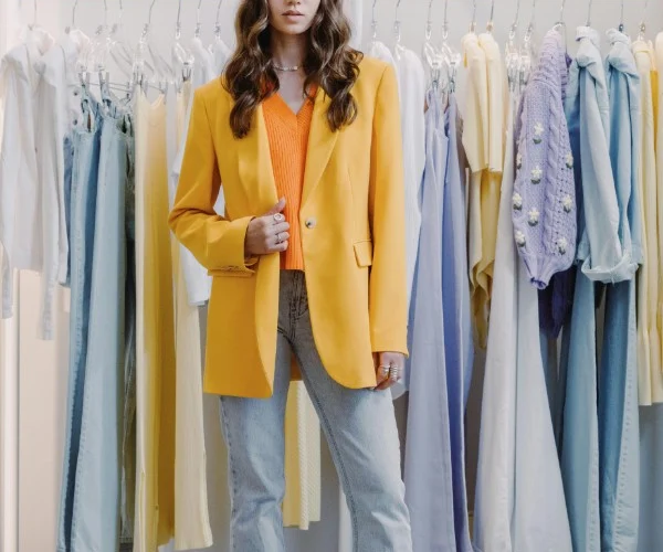 Ragazza indossa un outfit con blazer oversize giallo, top arancione e jeans chiari davanti a un appendiabiti, per la guida su come indossare i capi del guardaroba femminile.