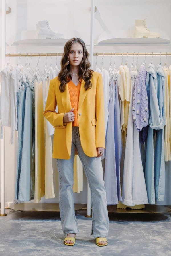 Ragazza indossa un outfit con blazer oversize giallo, top arancione e jeans chiari davanti a un appendiabiti, per la guida su come indossare i capi del guardaroba femminile.