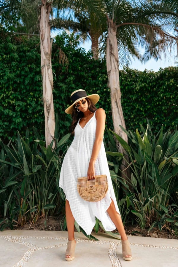 Donna indossa outfit per il mare: caftano con orlo asimmetrico bianco, cappello di paglia con nastro nero, borsa in paglia, occhiali da sole e sandali