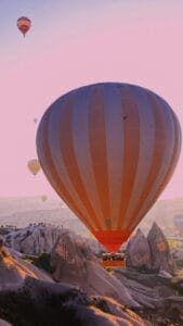 ​Una grande mongolfiera in volo sopra le formazioni rocciose al tramonto, con altre mongolfiere più piccole visibili sullo sfondo