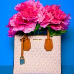 Borsa a mano Michael Kors con motivo logato bianco e dettagli in pelle marrone, riempita con grandi fiori fucsia su sfondo azzurro.