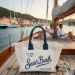 Borse e pochette Saint Barth: modello tote bag in canvas con frange sul ponte di una barca a vela con porticciolo e case sullo sfondo.
