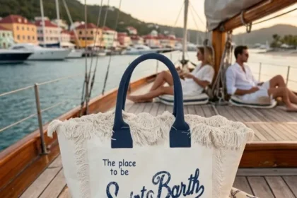 ​Borse e pochette Saint Barth: modello tote bag in canvas con frange sul ponte di una barca a vela con porticciolo e case sullo sfondo.