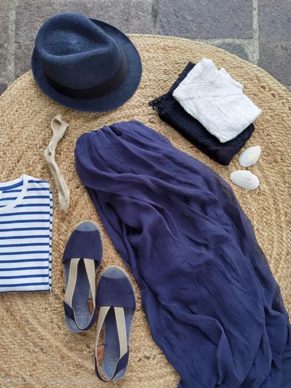 Flat lay di outfit estivo con colori tipici dello stile marinaro su tappeto di iuta con gonna fluida blu navy, espadrillas, cappello in paglia blu, maglietta piegata a righe bianche e blu, top pizzo e conchiglie decorative