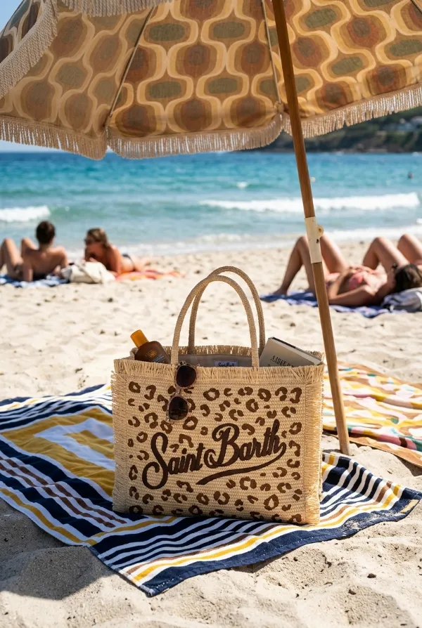 Borsa MC2 Saint Barth con stampa leopardata appoggiata su un telo mare a righe sulla sabbia, all'ombra di un ombrellone con motivo geometrico. Sullo sfondo, persone che prendono il sole e il mare cristallino.