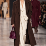 Modella in passerella con i colori moda Autunno Inverno 2026-2027: cappotto marrone cioccolato, tailleur crema e borsa berry