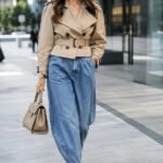 Donna con jeans balloon, trench corto stretto in vita e décolleté con tacco in un outfit metropolitano.
