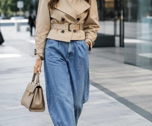 Donna con jeans balloon, trench corto stretto in vita e décolleté con tacco in un outfit metropolitano.